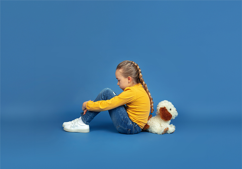 Portrait of sad little girl sitting isolated on blue studio background. How it feels to be autist. Modern problems, new vision of social issues. Concept of autism, childhood, healthcare, medicine.