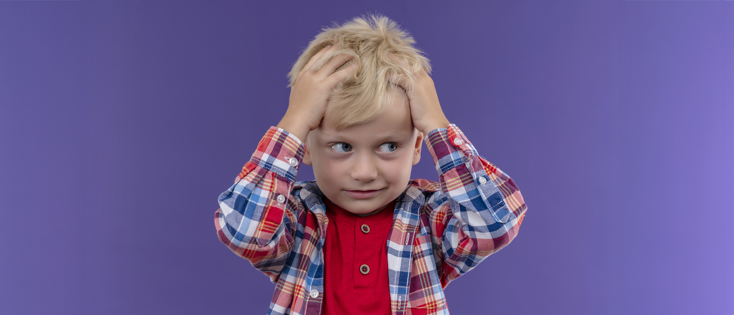 a cute little boy with blonde hair wearing checked shirt keeping hand on his head on a purple background