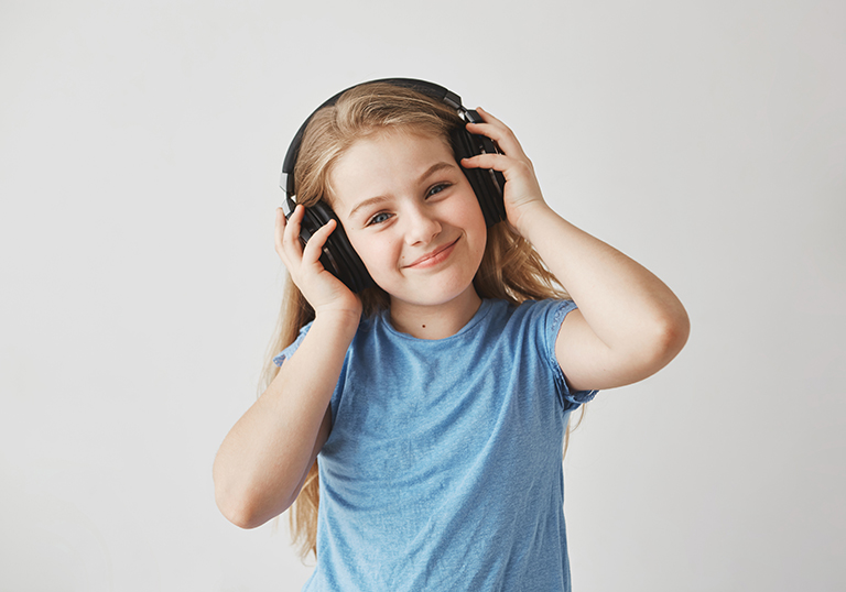 Portrait of cheerful little girl with light long hair and blue eyes in big earphones, listening to music with happy expression